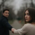 Женщина размышляет о прошлом на фоне расплывчатых фигур пары в парке.